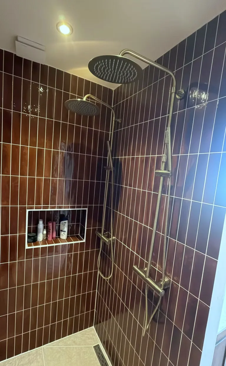 De afgeronde douche van Maj-Britt met donkerrode tegels, een messing regendouche en een betegelde nis