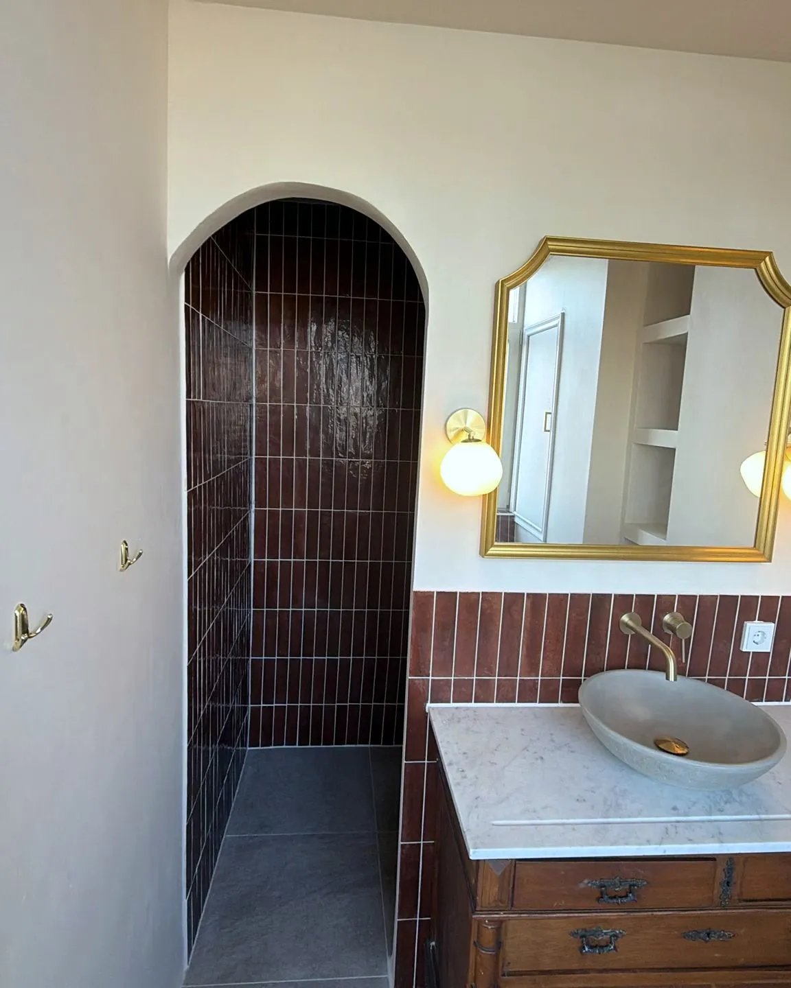 Dezelfde badkamer afgerond: donkerrode tegels, een boogvormige doorgang naar de douche en een houten wastafelmeubel