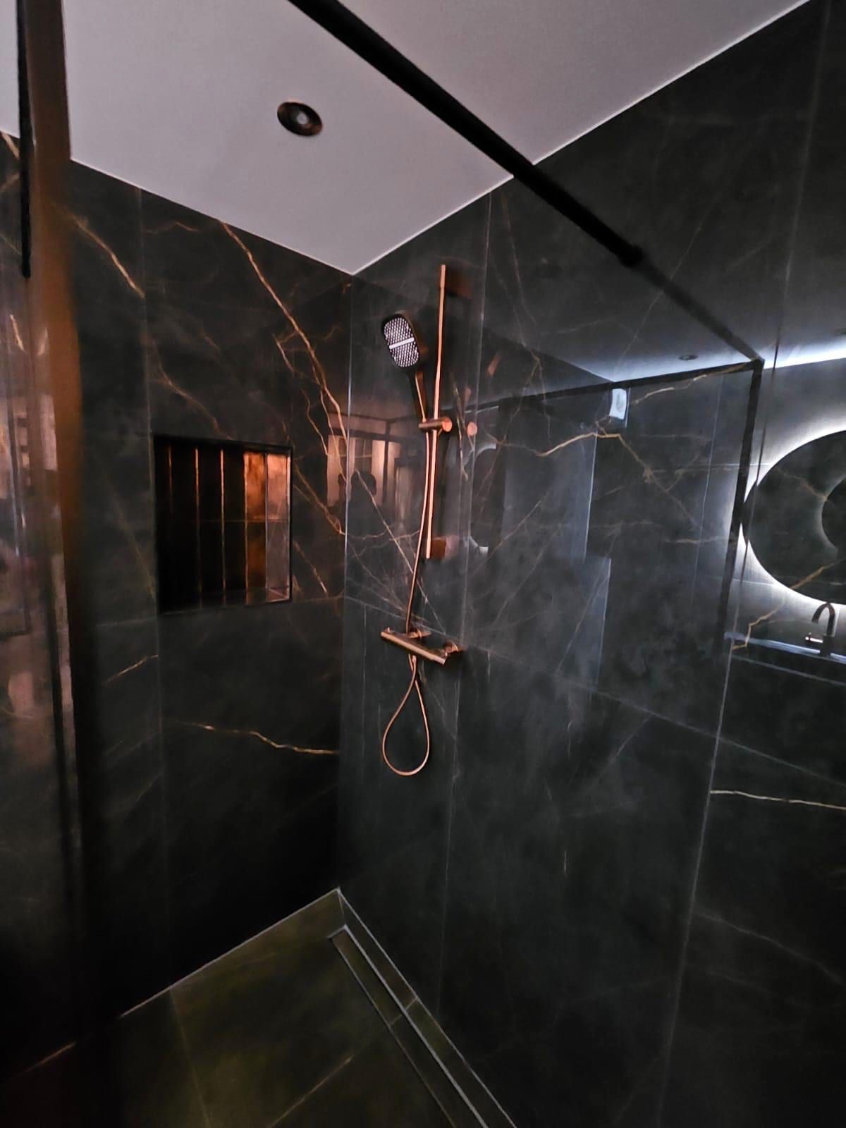 Koperkleurige doucheset tegen een wand van donkere marmerlook, met een glazen wand en een verlichte nis links