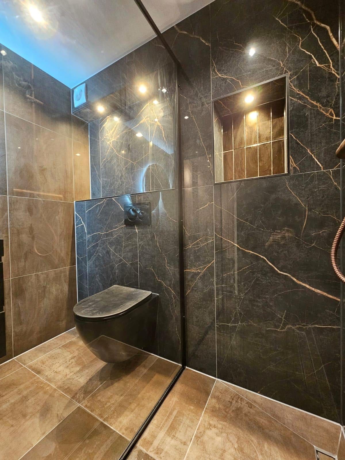 Zwevend toilet in mat antraciet tegen een wand van donkere marmerlook met goudkleurige adering, met een verlichte nis in goudkleurige tegel ernaast