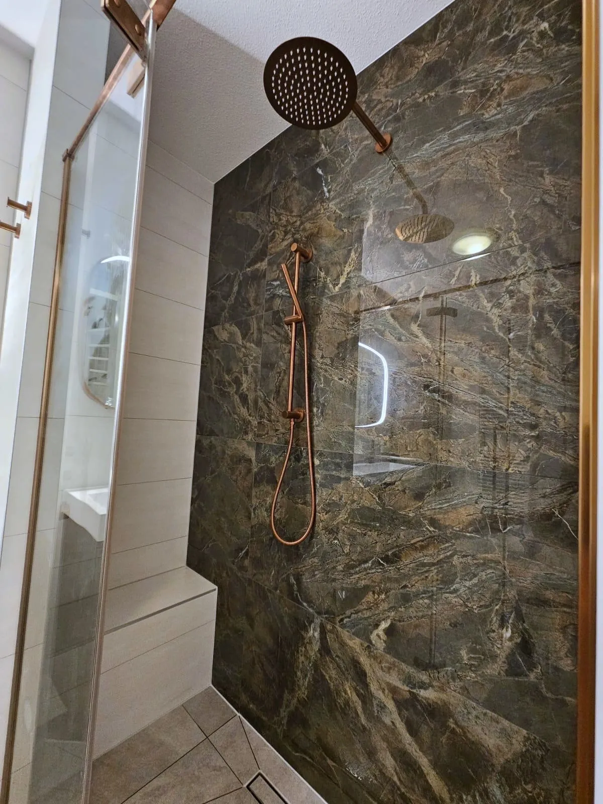 Inloopdouche met een wand van groenbruine marmerlook tegels, een koperkleurige regendouche en handdouche, een zitbank en een glazen wand met een koperkleurig profiel