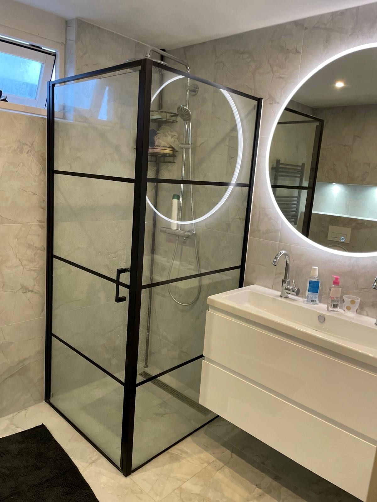 Badkamer met een zwart omlijste douchecabine, een ronde spiegel met verlichting en een wit hoogglans wastafelmeubel tegen grijze marmerlook