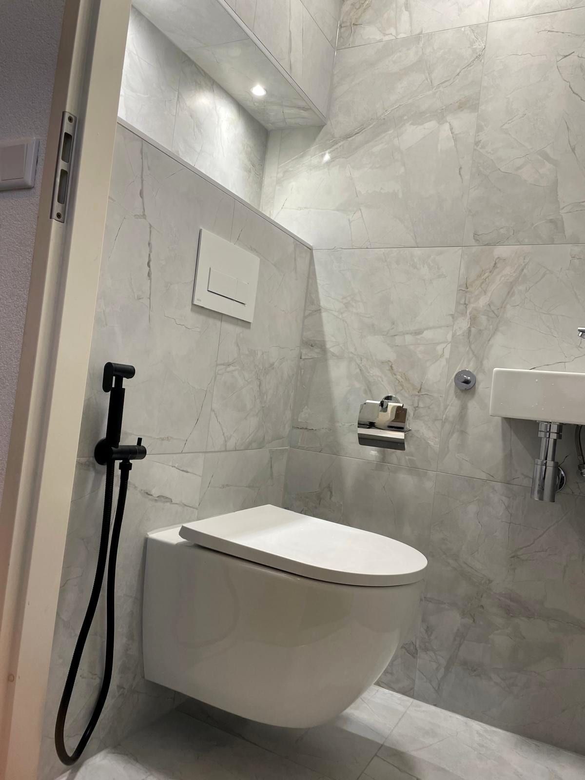 Zwevend toilet met een verlichte schapnis erboven, een zwarte bidetkraan ernaast en een kleine wastafel rechts