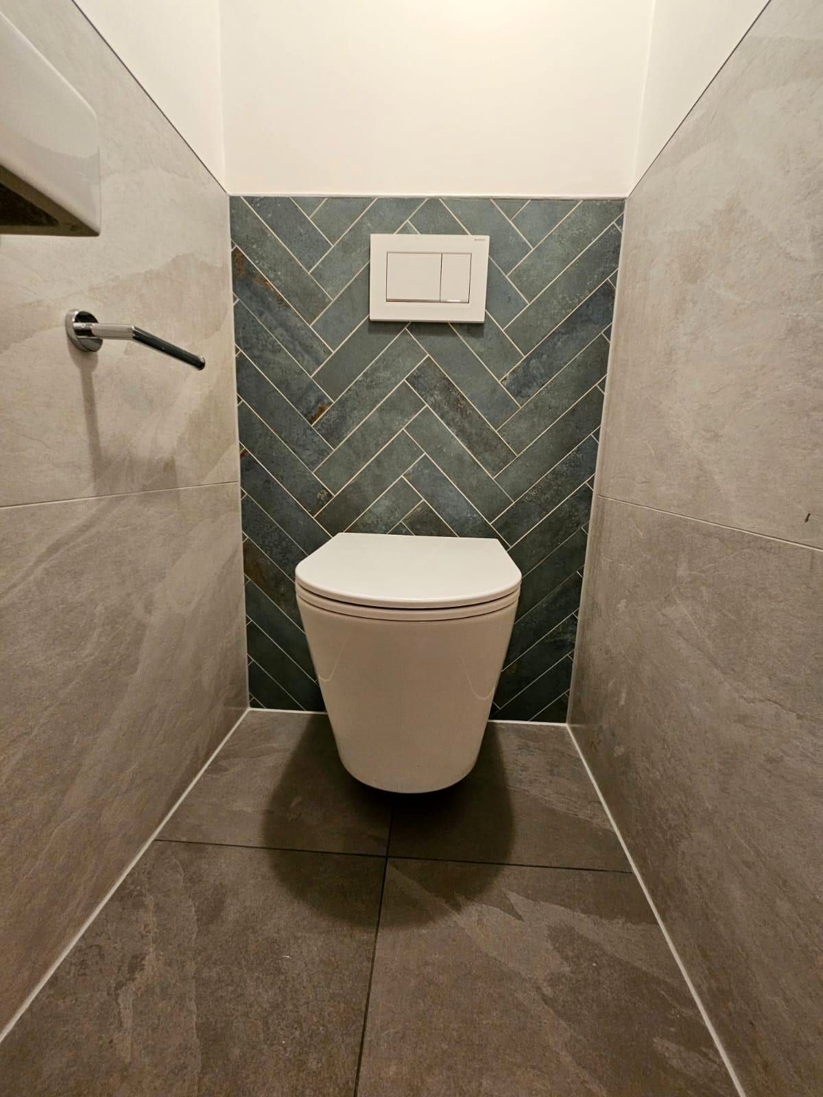 Zwevend toilet tegen een paneel van groene visgraattegel, met warme steenlook tegels ernaast en een witte bedieningsplaat