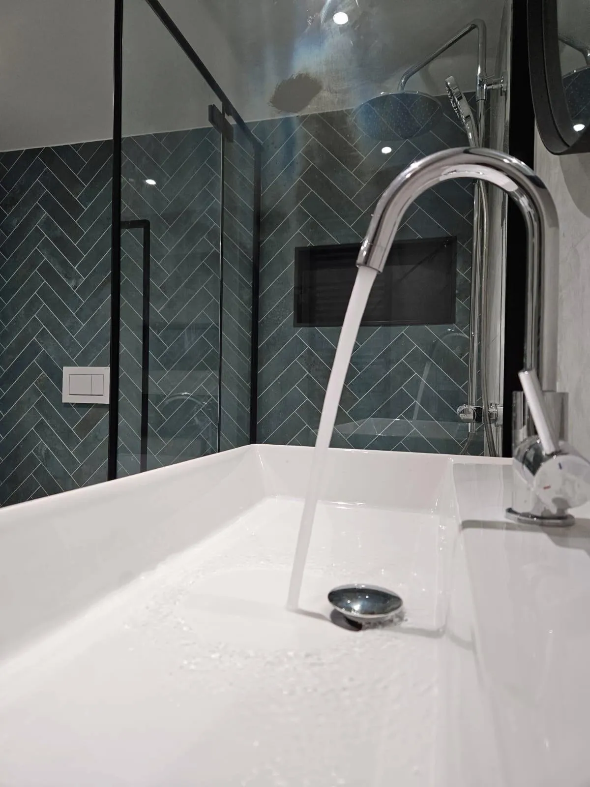 Chromen kraan met stromend water in een witte wasbak, met op de achtergrond een douchewand van groene visgraattegels achter glas