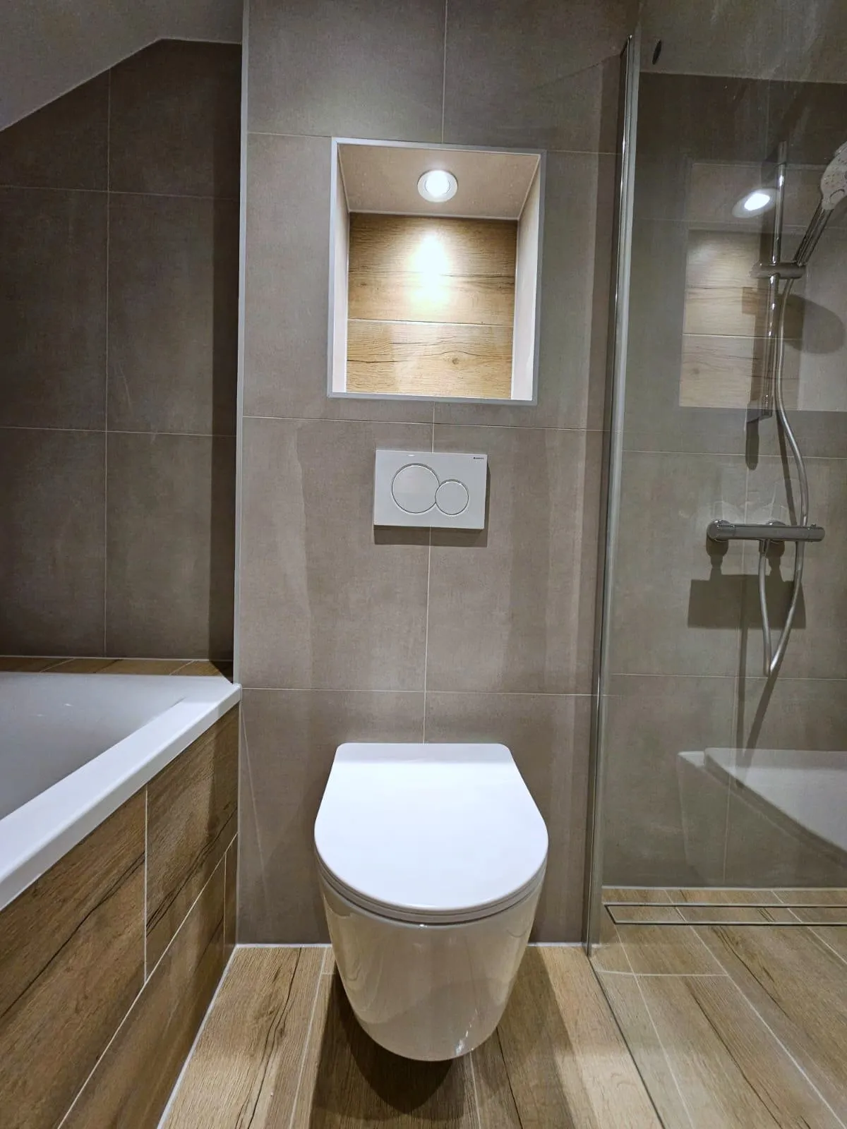 Badkamer met een ligbad met een ombouw in houtlook, een zwevend toilet onder een verlichte nis met houtlook tegels en een douche achter glas, op een vloer in houtlook