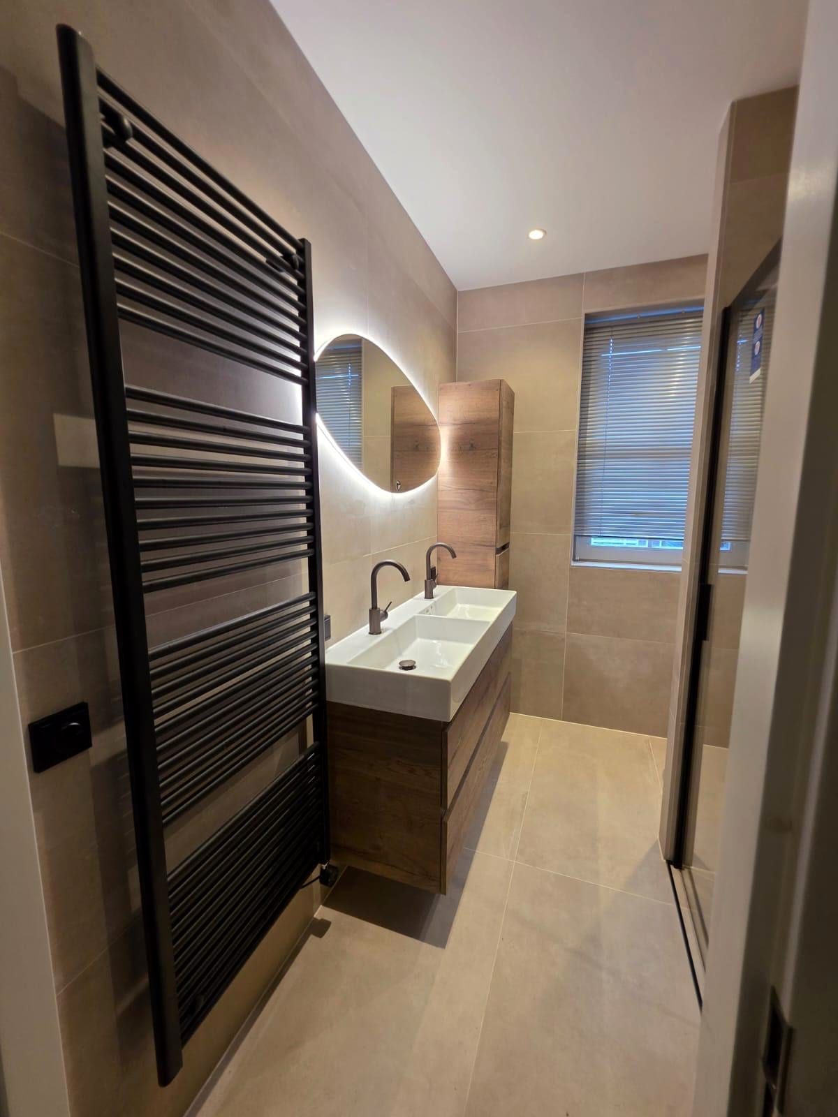 Badkamer met grote travertijnlook tegels, een zwarte handdoekradiator over de volle hoogte, een ovale spiegel met verlichting en een wastafelmeubel in houtlook met een brede witte wastafel