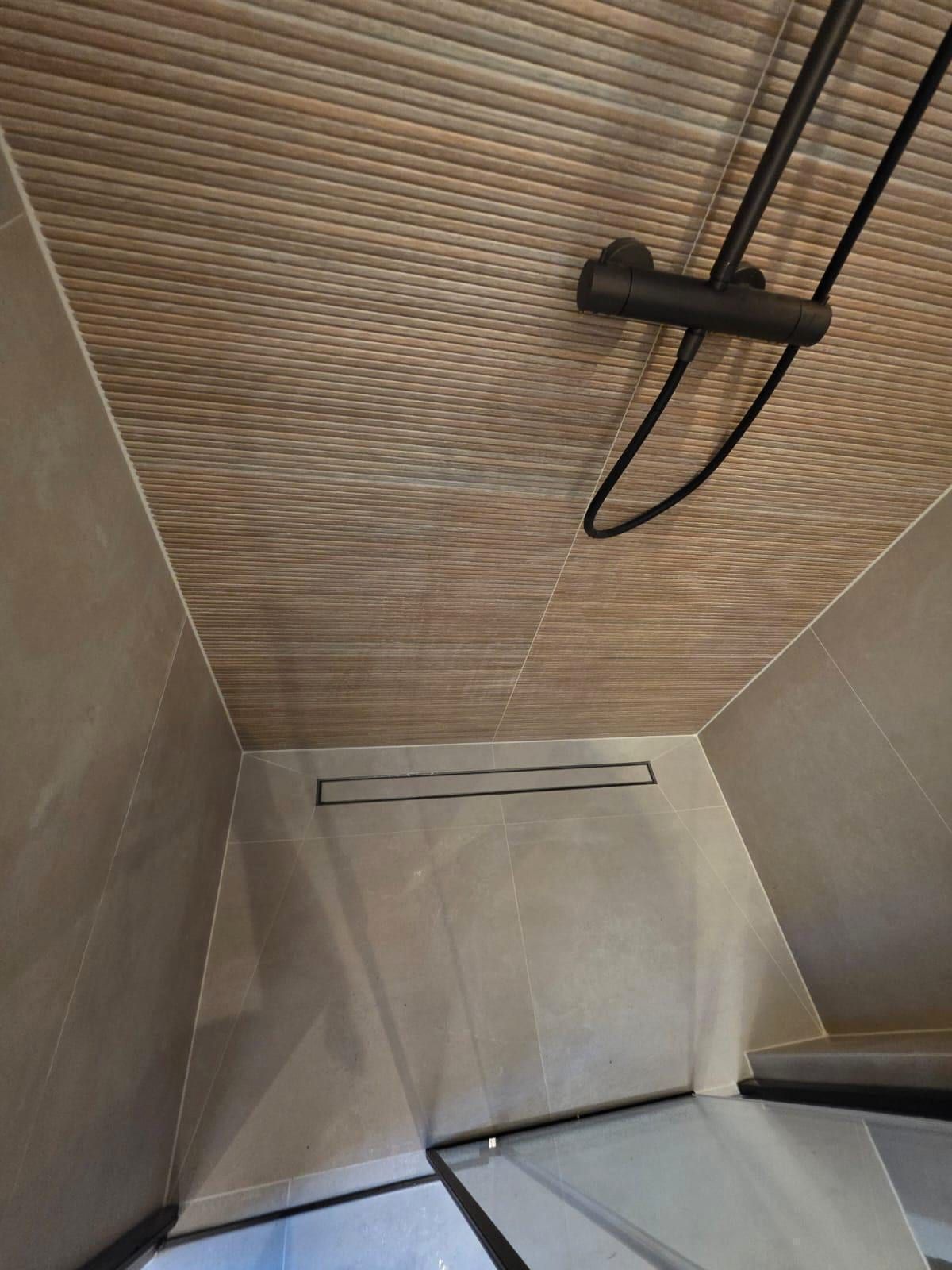 Douchevloer met een lange donkere afvoergoot, doorlopende travertijnlook tegels en een zwarte thermostaatkraan aan de ribbelwand in houtlook