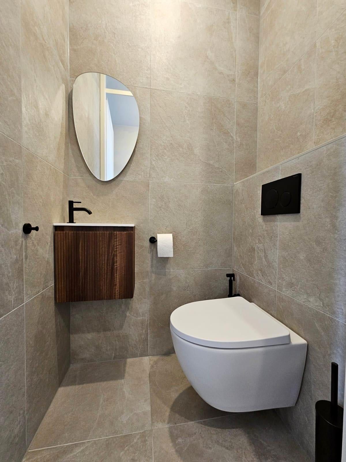 Apart toilet met een klein notenhouten wastafelmeubel, een zwarte kraan en een spiegel in een organische vorm