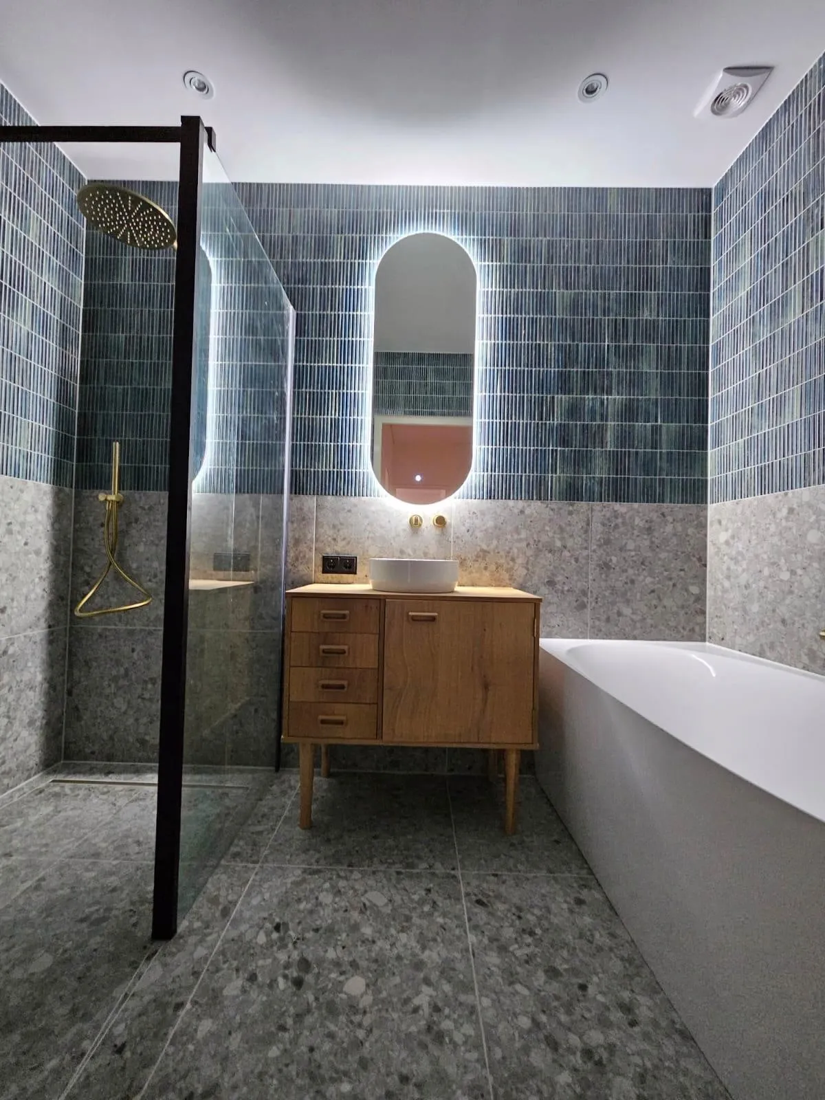 Badkamer met een terrazzo vloer, blauwe staafjestegels op de wanden, een eiken wastafelmeubel met een opzetkom onder een ovale verlichte spiegel, een ligbad en een inloopdouche met zwart profiel