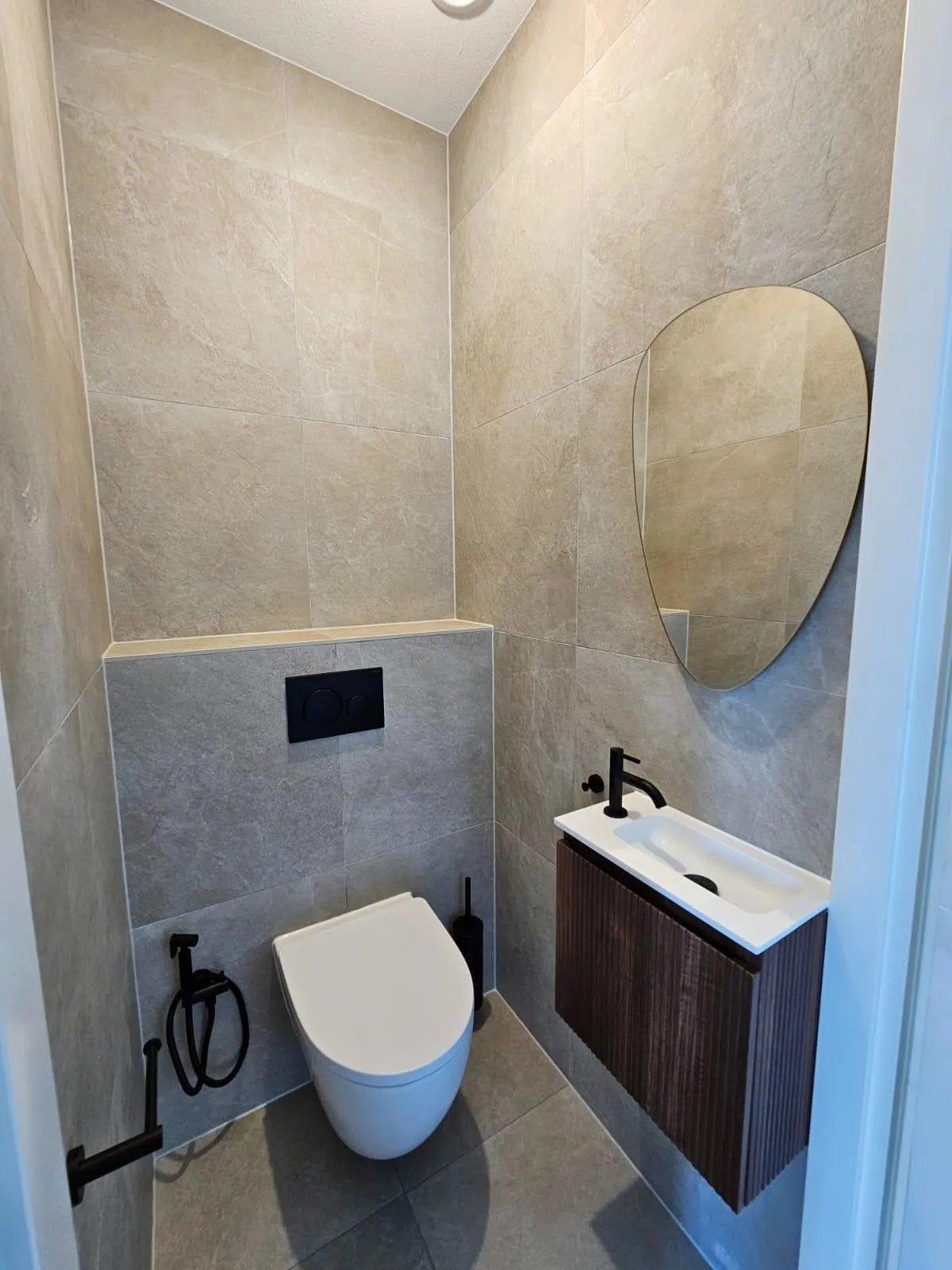 Toiletruimte met een notenhouten fonteinmeubel met een zwarte kraan, een zwart bedieningspaneel, een bidetsproeier naast het zwevende toilet en een organisch gevormde spiegel