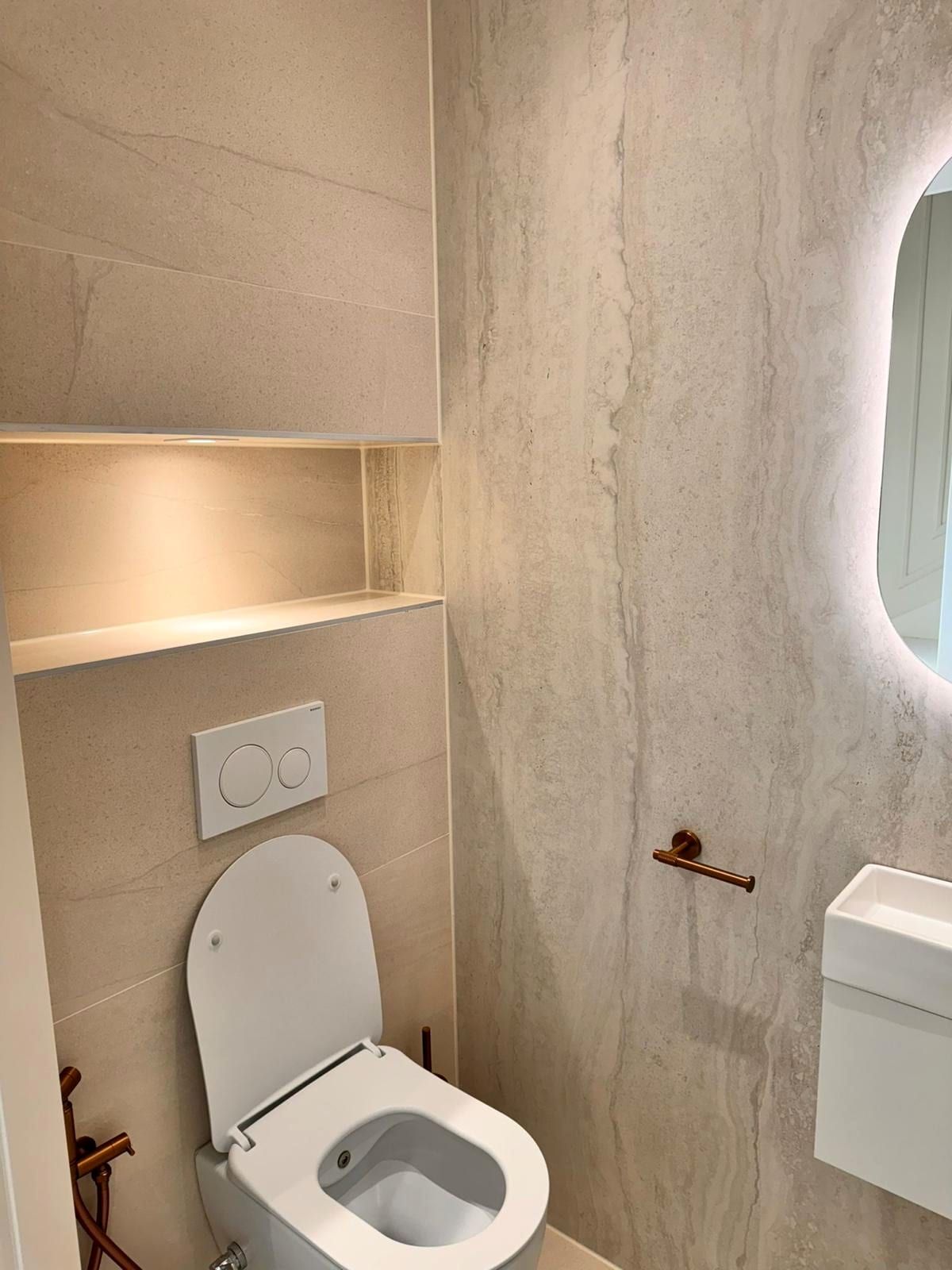 Zwevend toilet met een verlichte schapnis erboven, een koperkleurige rolhouder en een spiegel met verlichting in een organische vorm ernaast