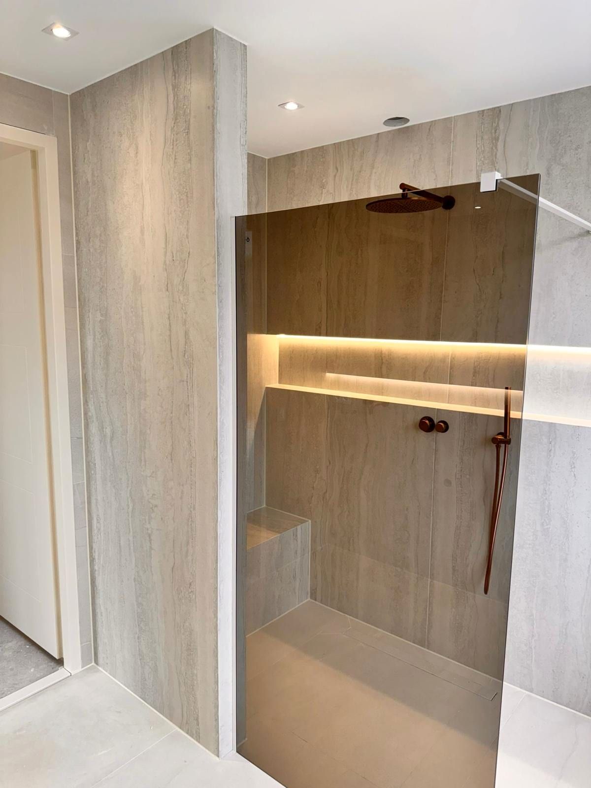 Douche met een koperkleurige regendouche aan een wandarm, een gemetselde zitbank en een verlichte nis in de wand