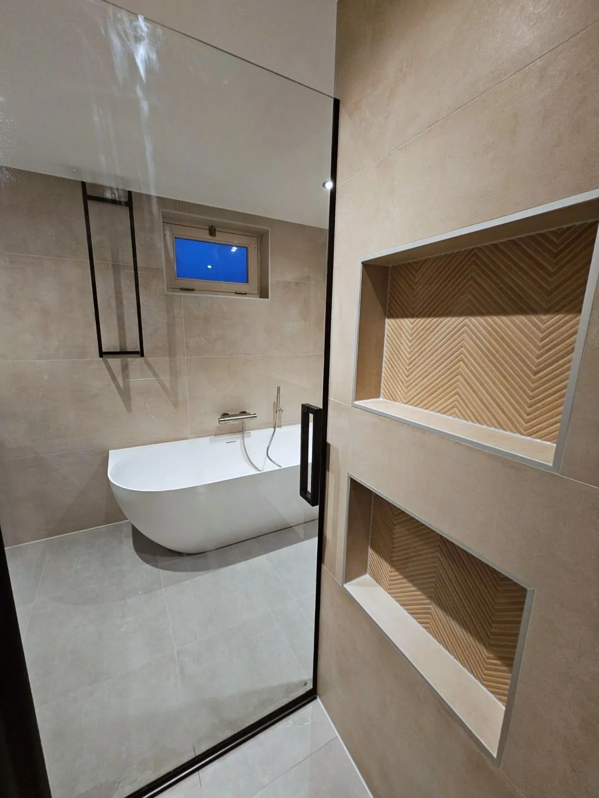 Badkamer met een vrijstaand bad bij een klein raam, gezien door een glazen douchewand met zwart profiel, en twee nissen met houtlook visgraattegels in de wand