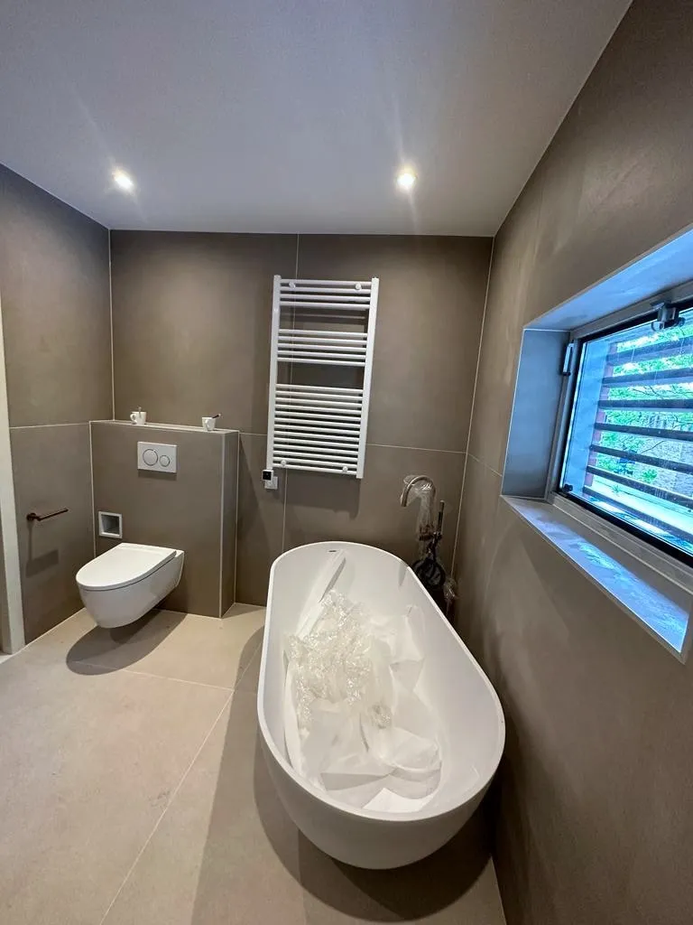 Badkamer met een vrijstaand bad onder een langwerpig raam, een zwevend toilet en een witte handdoekradiator