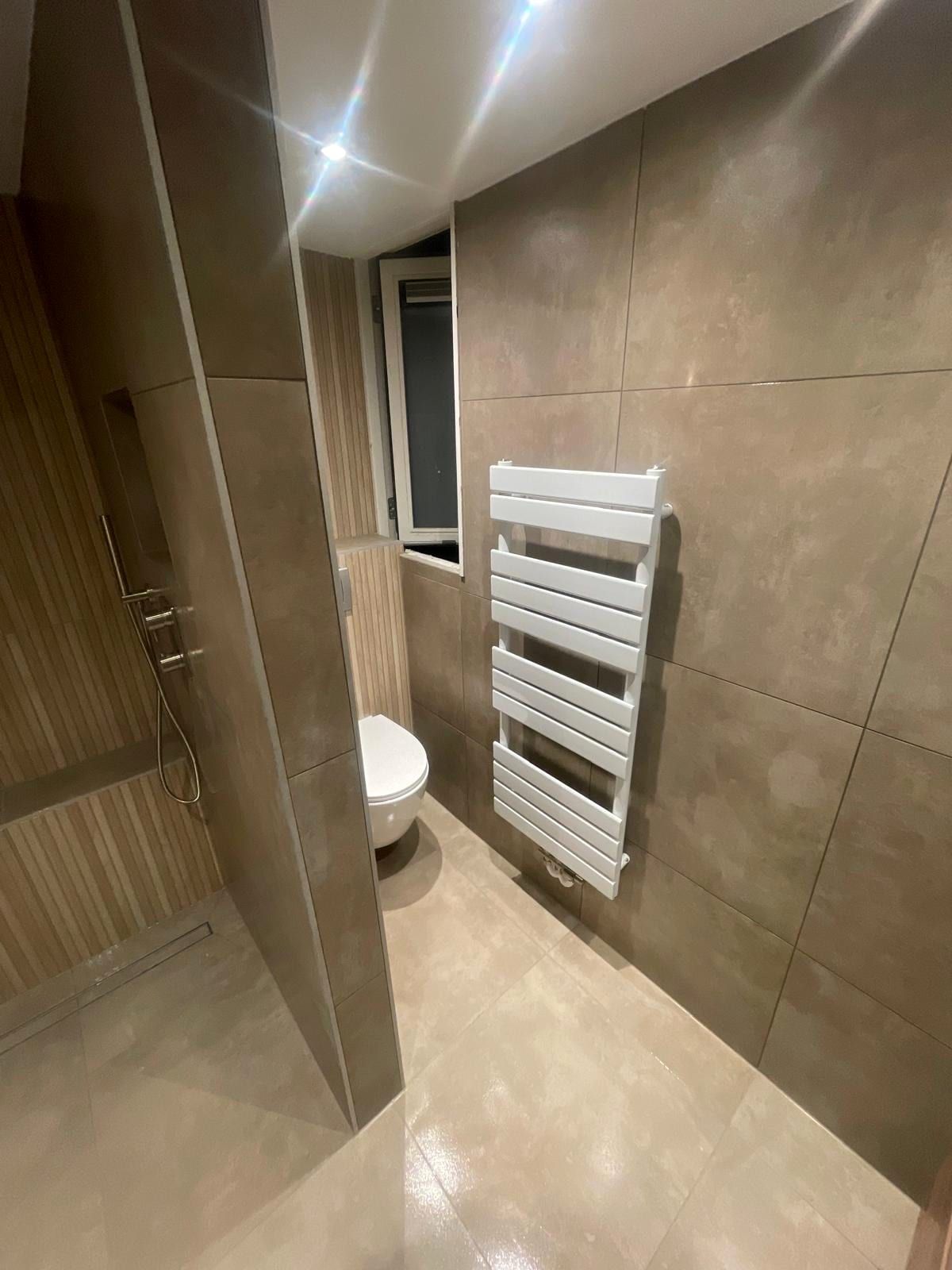 Zwevend toilet achter een gemetselde wand, met een witte handdoekradiator en een raam ernaast
