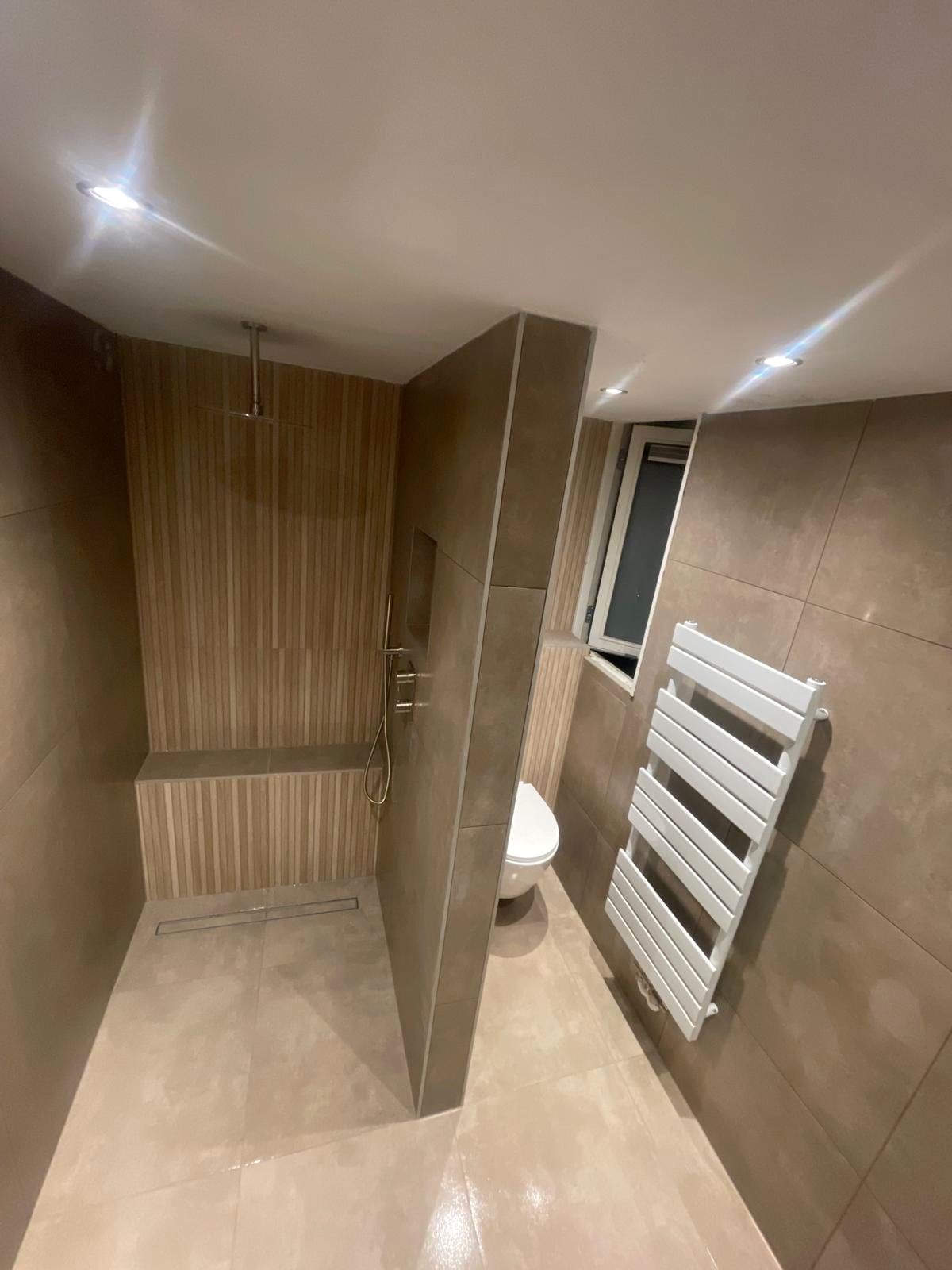 Badkamer met een ribbelwand in houtlook in de doucheruimte, een regendouche aan het plafond en een witte handdoekradiator bij het raam
