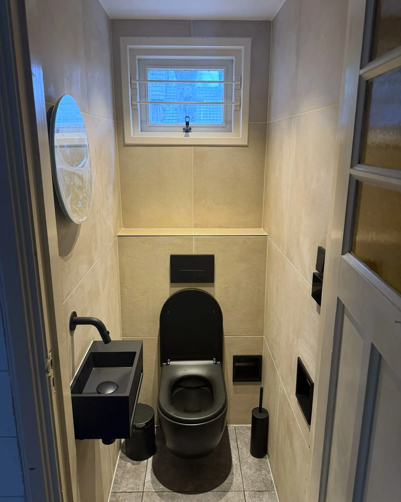 Hetzelfde toilet na de renovatie: grote lichte tegels, een zwevende zwarte pot met zwart bedieningspaneel, een zwarte fontein met wandkraan en nissen in het tegelwerk