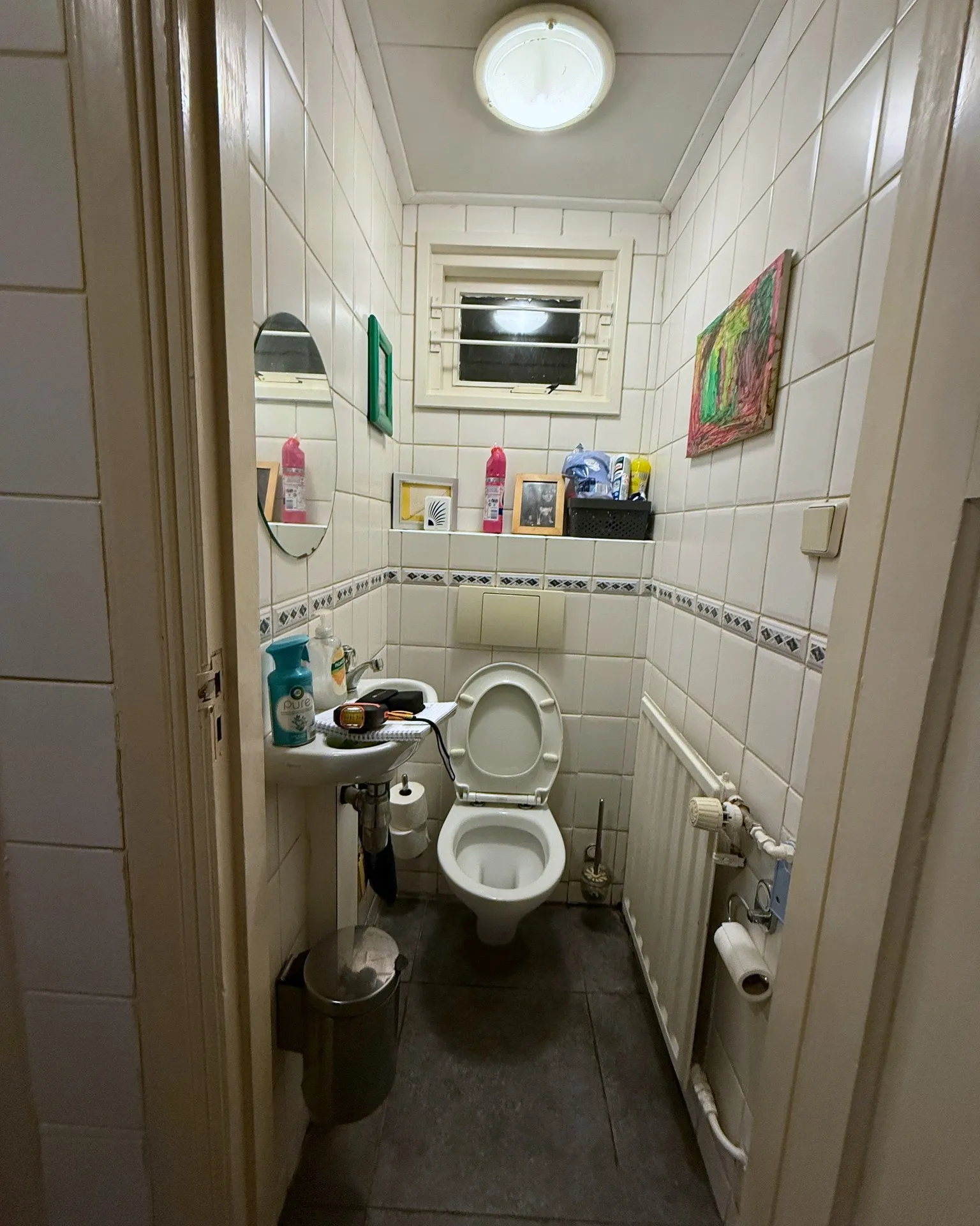 Een gedateerd toilet met witte tegels, een sierrand, een oude pot en een fontein met zichtbaar leidingwerk, voor de renovatie