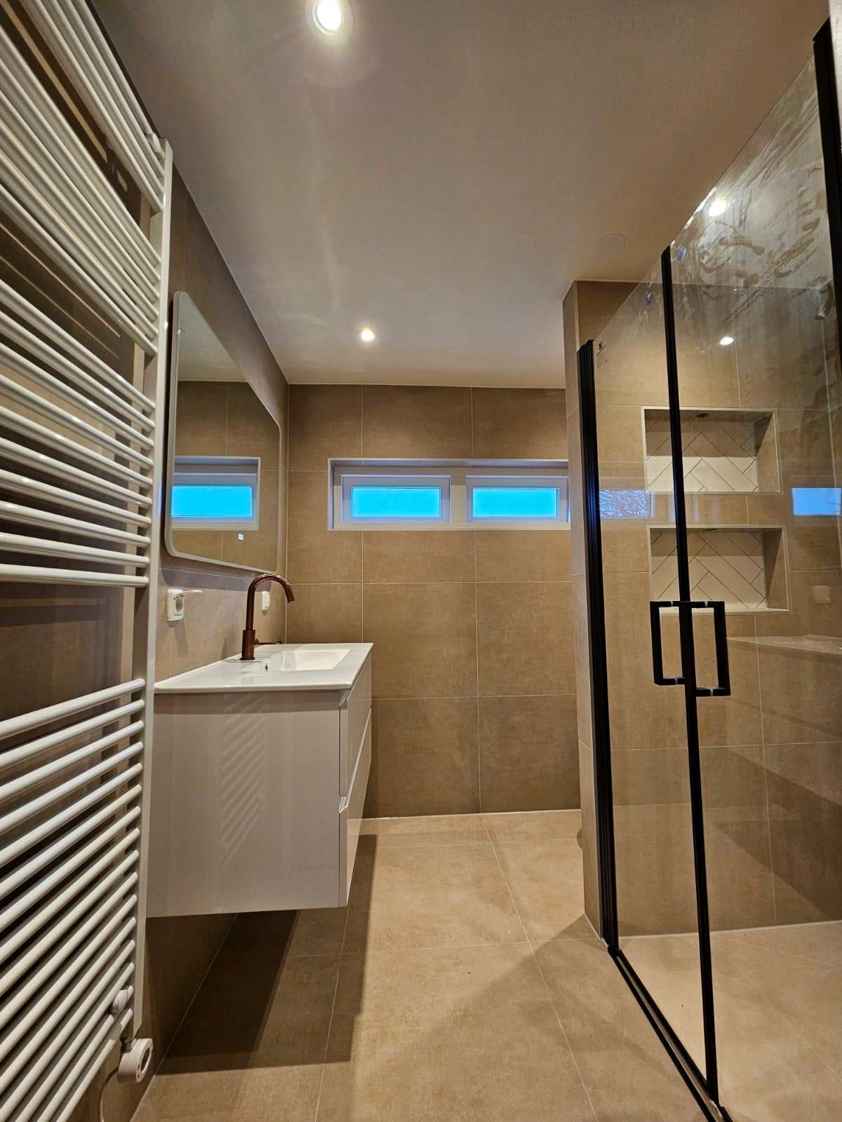 Badkamer met een wit hoogglans wastafelmeubel, een koperkleurige kraan, een hoge witte handdoekradiator en een zwart omlijste douchecabine