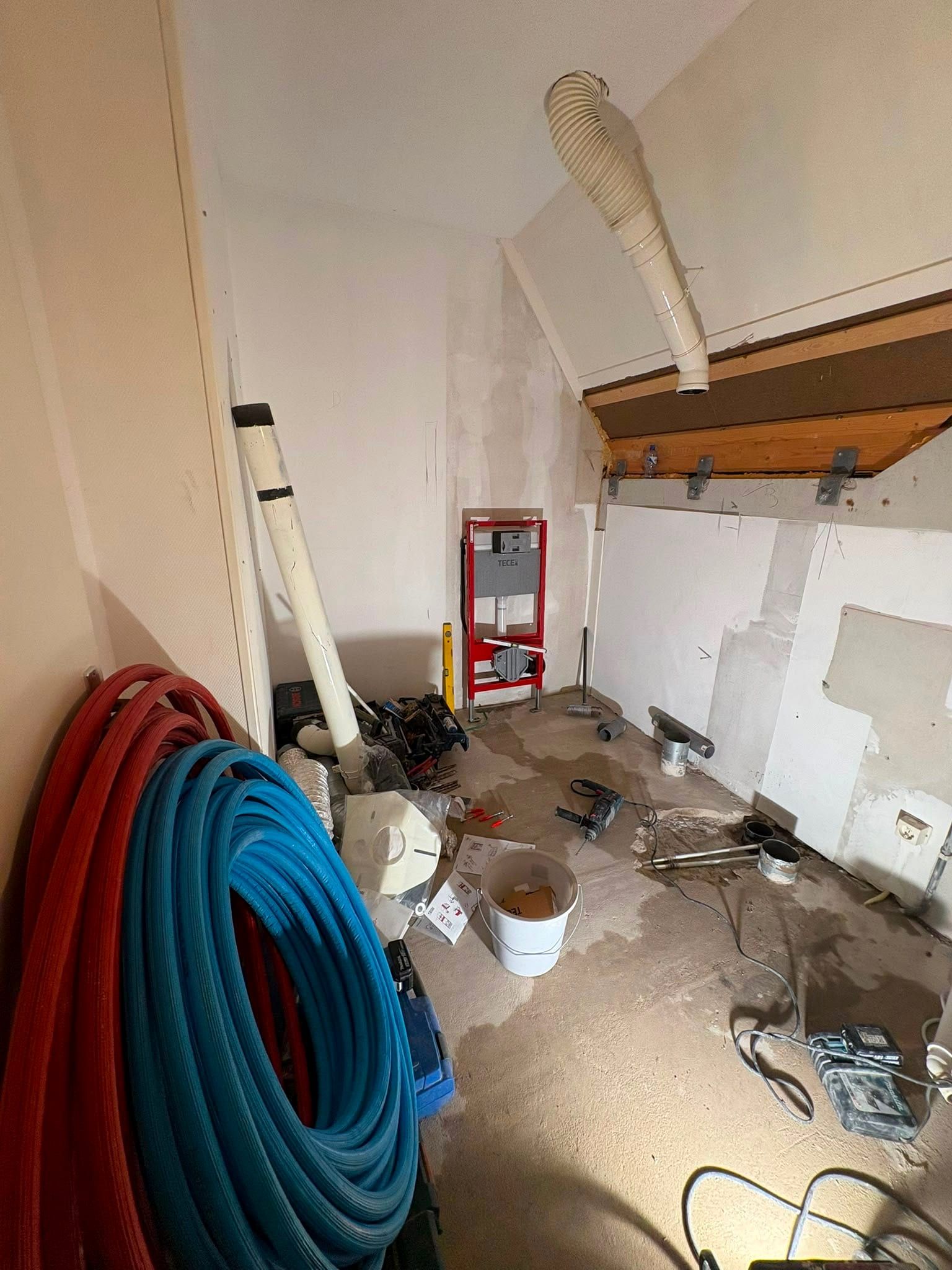Kale zolderruimte tijdens de verbouwing, met losliggende leidingen, een montageframe voor het toilet en bouwmateriaal op de vloer