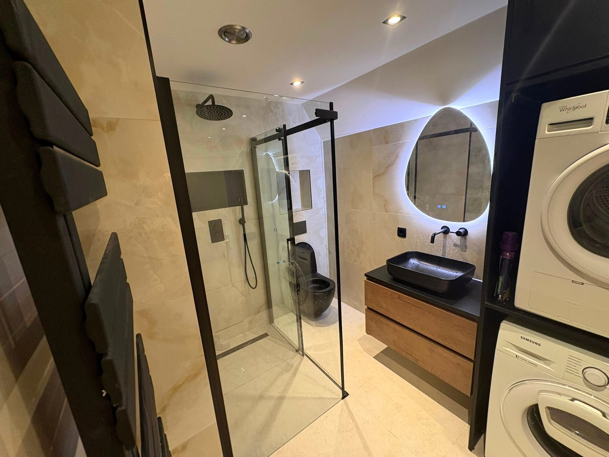 De afgeronde zolderbadkamer over de volle lengte, met de doucheruimte, het zwarte toilet, het wastafelmeubel in houtlook en de wasmachine met droger erop onder het schuine dak