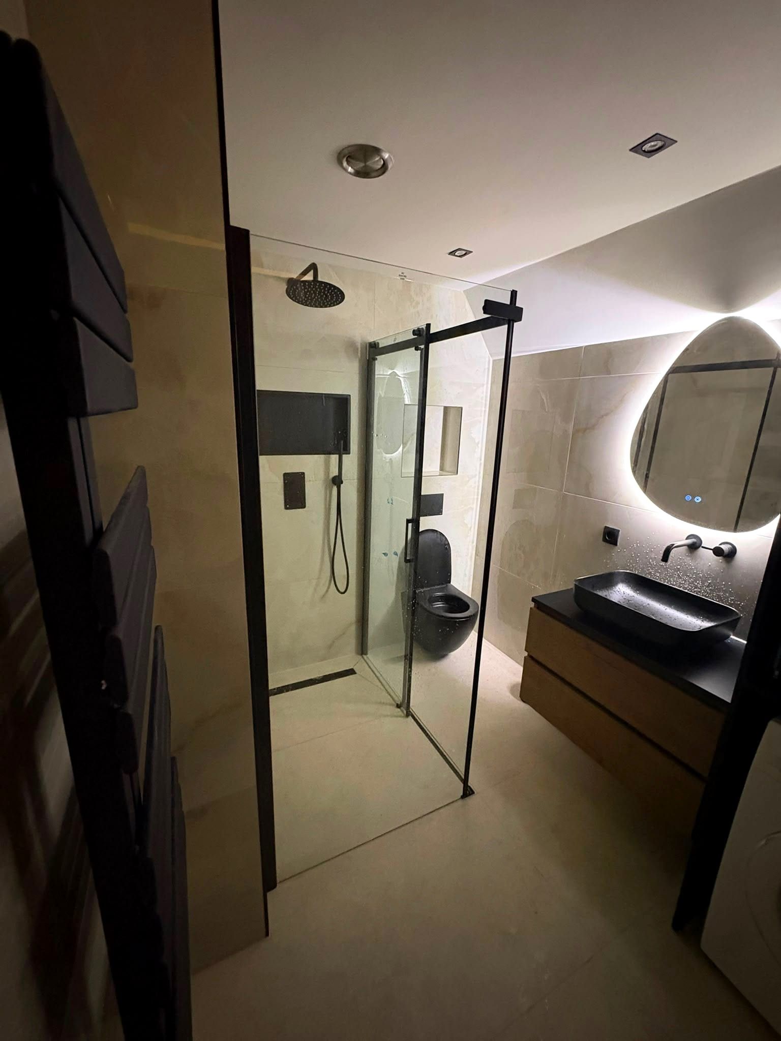 Afgeronde zolderbadkamer met een douche met zwart omlijste schuifdeur, een zwart zwevend toilet en een wastafelmeubel in houtlook met een zwart blad en een zwarte waskom